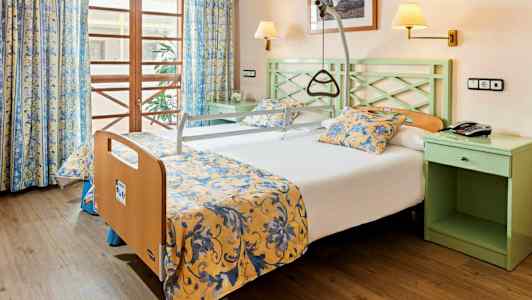 Hotelværelse med en seng udstyret med støttehåndtag og gul- og blomstermønstrede sengetæpper. (AI-genereret tekst)