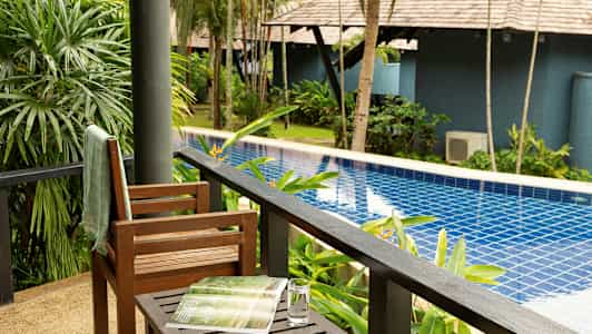 Udsigt fra en veranda mod en pool omgivet af grønne planter og træer. (AI-genereret tekst)