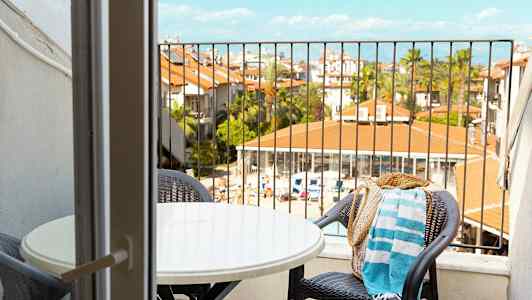 Udsigt fra en altan med et bord og stole mod et poolområde og bygninger. (AI-genereret tekst)