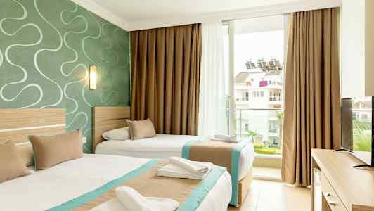 Et hotelværelse med to senge. Sengene er redt med hvide lagner og har beige og turkise sengetæpper. Værelset har en grøn væg med et mønster og tilbyder udsigt til en bygning uden for vinduet. (AI-genereret tekst)