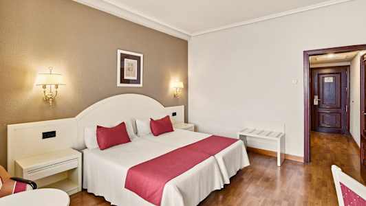 Et hotelværelse med en dobbeltseng med røde detaljer, væglamper og en dør i baggrunden. (AI-genereret tekst)