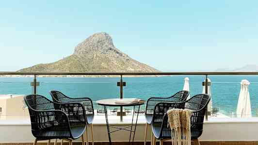 Fire stole og et bord på en terrasse med udsigt over havet og en bjergtop. (AI-genereret tekst)