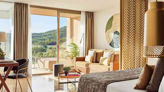 Et hotelværelse med en seng, siddeområde og udsigt over grøn natur gennem glasdøre. (AI-genereret tekst)