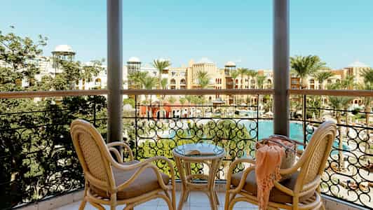 Udsigt fra en altan med to rottingsstole og et bord, mod en pool og hotel omgivet af palmer. (AI-genereret tekst)