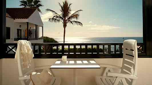 Udsigt fra en terrasse med bord og stole mod havet, med en palme og en bygning med rødt tag i baggrunden. (AI-genereret tekst)