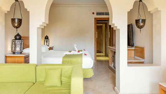 Et hotelværelse med en seng dekoreret med et formet håndklæde og blomster, en grøn sofa i forgrunden og udsmykkede lamper. (AI-genereret tekst)