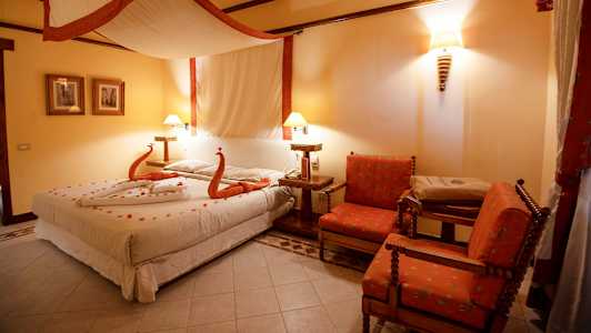 Et soveværelse med en seng dekoreret med håndklæder formet som svaner og kronblade, to røde lænestole og varm belysning. (AI-genereret tekst)