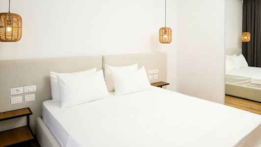 Et moderne hotelværelse med en stor seng redt med hvide lagner. Hængende lamper i rattan stil flankerer sengen. (AI-genereret tekst)