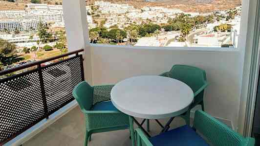 Udsigt fra en altan med et rundt bord og fire grønne stole, med udsigt over en by og bjerge i baggrunden. (AI-genereret tekst)