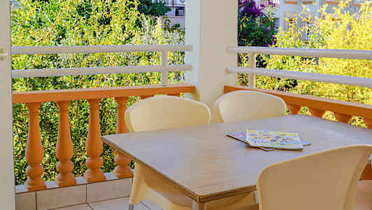 Billede af en altan med et bord og stole på flisebelagt gulv samt udsigt til beplantning og bygninger. (AI-genereret tekst)