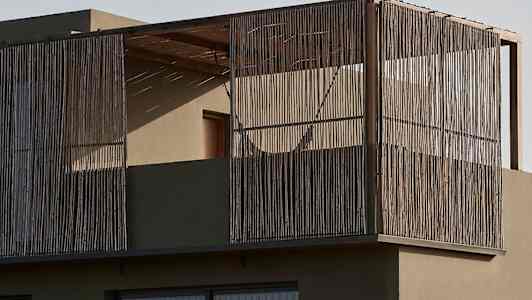 En moderne bygning med en altan omgivet af træstænger og glasvinduer. Foran ses vegetation i et ørkenlandskab. (AI-genereret tekst)