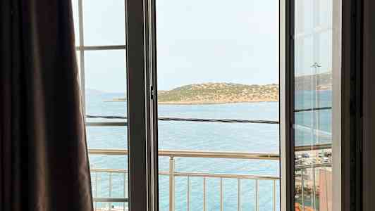 Udsigt fra et åbent vindue med en altan, havet og en ø i baggrunden. (AI-genereret tekst)