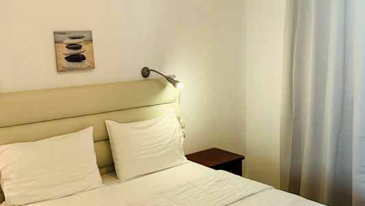 En redt seng med hvide sengetøj og to foldede håndklæder. Væggdekoration og gardiner i baggrunden. (AI-genereret tekst)