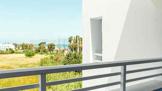 Udsigt fra en balkon med bord og stol, hvide bygninger og grønne områder i baggrunden. (AI-genereret tekst)