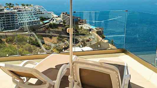 To liggestole under en parasol på en terrasse med udsigt over havet og nærliggende hoteller. (AI-genereret tekst)