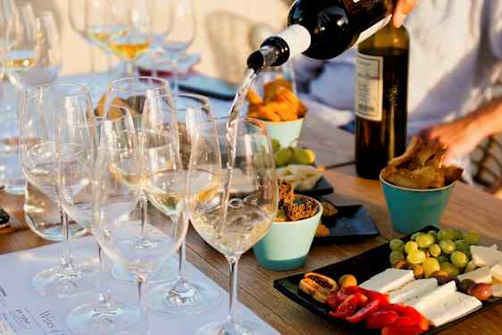 Et bord med glas fyldt med hvidvin, der serveres, omgivet af forskellige snacks som ost, brød og frugt. (AI-genereret tekst)