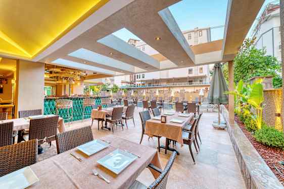 Udendørs restaurant med borde dækket med tallerkener og bestik, samt grønne planter og pool i baggrunden. (AI-genereret tekst)