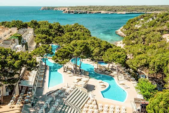 Udsigt over en ferieanlæg med swimmingpools omgivet af liggestole og parasoller, beliggende ved siden af et turkisblåt hav med frodig vegetation i baggrunden. (AI-genereret tekst)