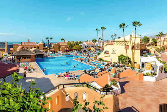 Et hotelområde med en stor pool omgivet af liggestole og bygninger. (AI-genereret tekst)