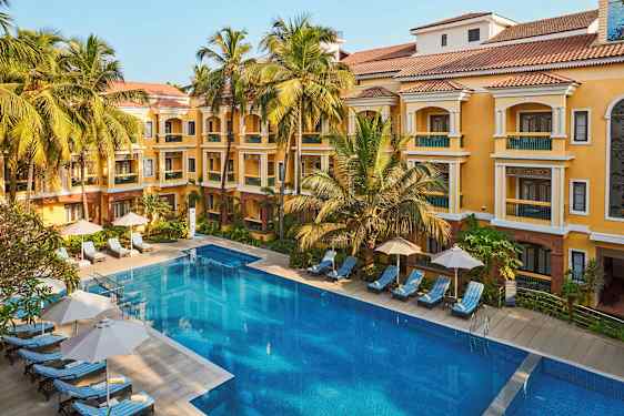 Et billede af en hotelbygning med gul facade og rødt tag, omgivet af palmer. Foran bygningen er der en stor swimmingpool og liggestole med parasoller. (AI-genereret tekst)