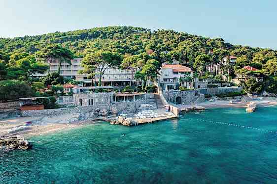 En bygning omgivet af grønne områder tæt på en strand med klart vand. (AI-genereret tekst)