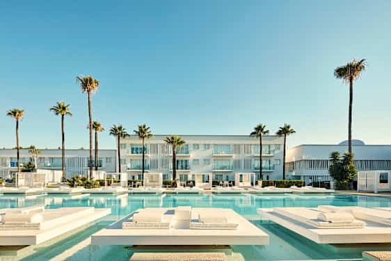 En moderne bygning med altaner, omgivet af palmer og en swimmingpool med liggestole. (AI-genereret tekst)