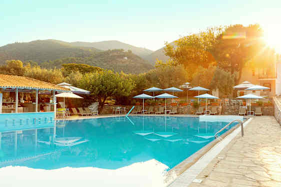 En udendørs pool med liggestole og parasoller, omgivet af træer og bygninger i et bjergrigt landskab. (AI-genereret tekst)