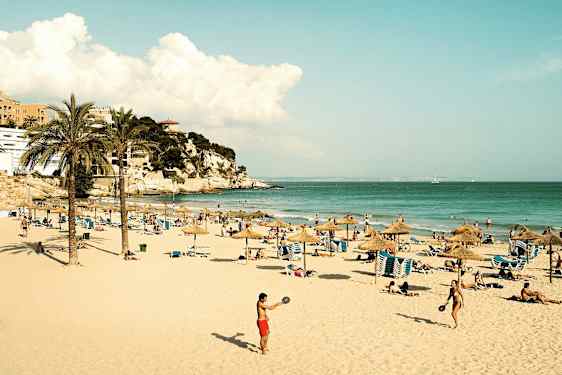 En sandstrand med parasoller, liggestole og mennesker, der nyder solen og havet. Længere væk ses en klippe med bygninger. (AI-genereret tekst)