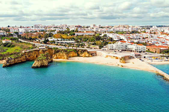 Lys sandstrand ved havet med en by i baggrunden og klipper ved stranden. (AI-genereret tekst)