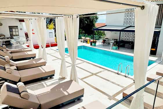 Udendørsbillede af en pool omgivet af solsenge og pergola med hvide gardiner. (AI-genereret tekst)