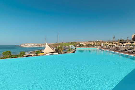 En stor udendørs pool med liggestole og parasoller ved siden af, med udsigt over havet og en kystby. (AI-genereret tekst)