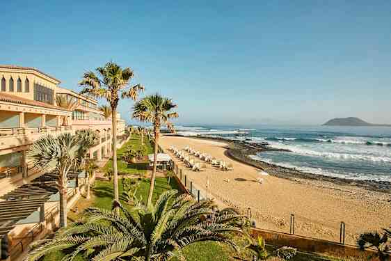 En strand med sand, parasoller og bølger, omgivet af bygninger og palmer. (AI-genereret tekst)