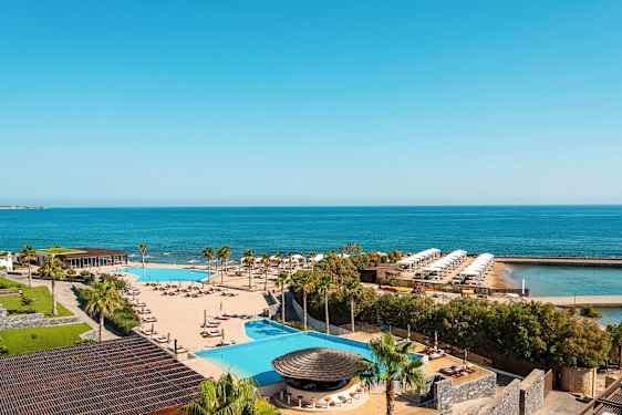 Udsigt over et resort med pool, solsenge, palmer og hav i baggrunden. (AI-genereret tekst)