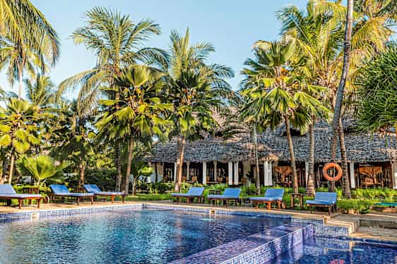 En pool omgivet af solsenge og palmer ved en bygning med stråtag. (AI-genereret tekst)