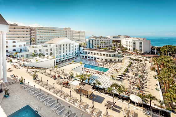 Udsigt over et hotelområde med pools, solsenge og hvid arkitektur tæt på havet. (AI-genereret tekst)