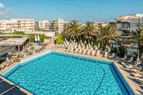 Billedet viser en udendørs swimmingpool med solsenge og parasoller, omgivet af palmer og bygninger. (AI-genereret tekst)