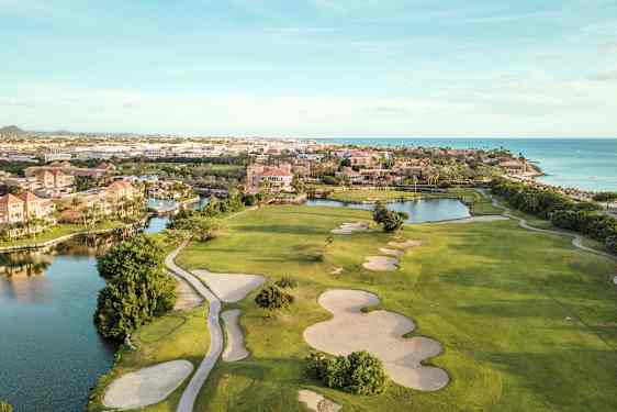 Golfbane med sandbunkere og grønne græsarealer nær vandløb og strand. Bebyggelse i baggrunden. (AI-genereret tekst)