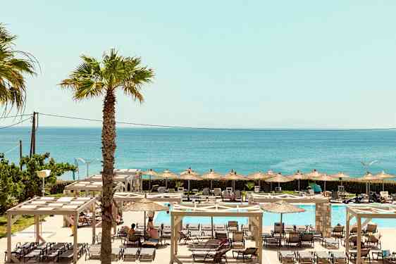 En udsigt over udendørs poolområde med liggestole, parasoller og havet i baggrunden. (AI-genereret tekst)