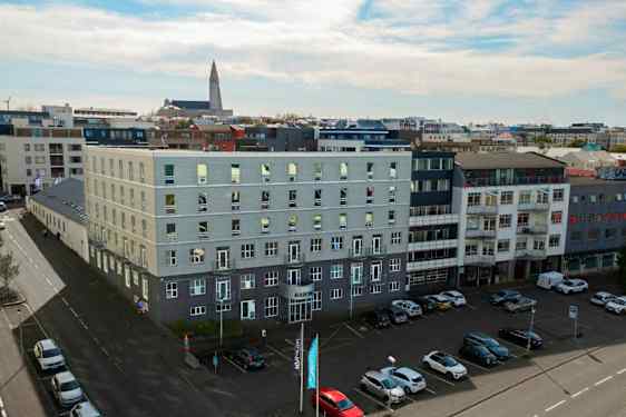 Flere moderne bygninger omgiver et åbent parkeringsområde, med biler parkeret på jorden og en kirke synlig i baggrunden. (AI-genereret tekst)