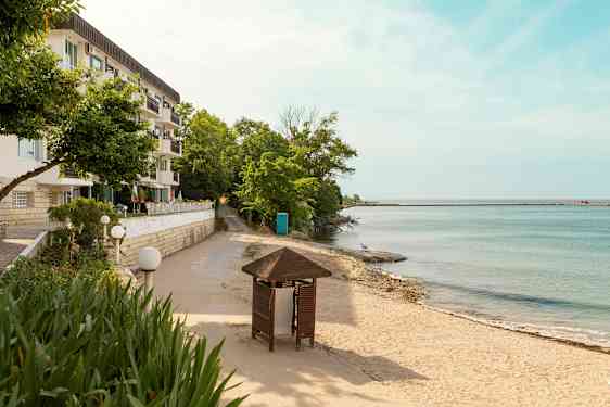 En bygning med flere altaner ved siden af en sandstrand og hav med træer og en pavillon i forgrunden. (AI-genereret tekst)