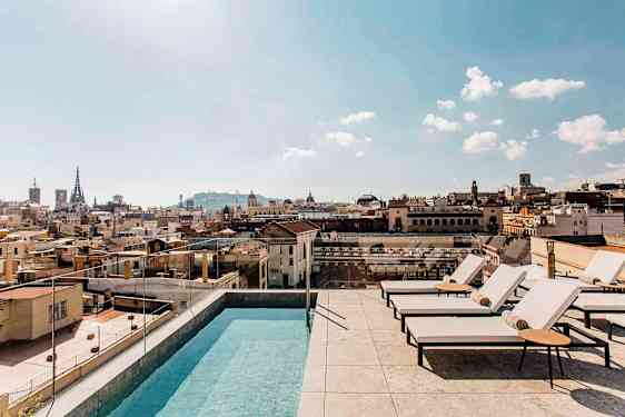 En udendørs pool og liggestole på en tagterrasse med udsigt over en by og dens bygninger under en klar himmel. (AI-genereret tekst)