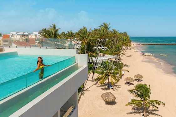 En swimmingpool med glasrækværk på en bygning med udsigt over en strand med palmer og asurblåt hav. (AI-genereret tekst)