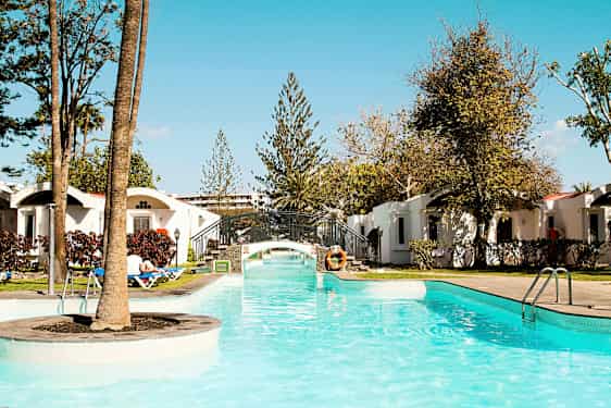 En pool med klart vand og en lille bro, omgivet af grønne områder og ferieboliger. (AI-genereret tekst)