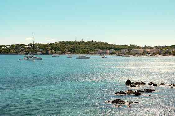 Udsigt over en kystlinje med klart blåt vand, flere både og sejlbåde i nærheden af en frodig strand og en by. (AI-genereret tekst)