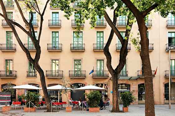 Et hotel med flere etager og altaner, et fortov med parasoller og siddepladser mellem træer. (AI-genereret tekst)