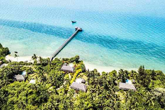 Luftfoto af en tropisk kystlinje med huse omgivet af frodig vegetation. En bro strækker sig ud i det turkise vand. (AI-genereret tekst)