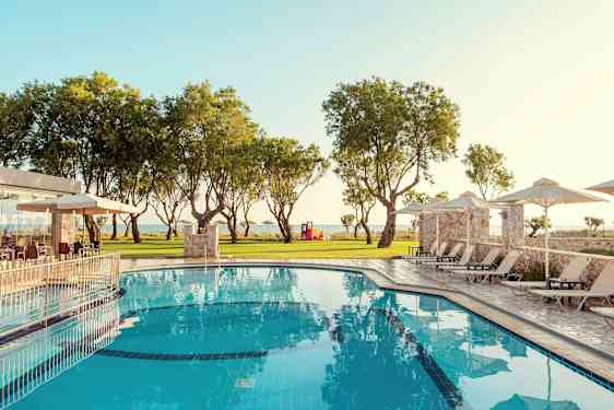 En pool med liggestole og parasoller omgivet af grønne områder og træer. (AI-genereret tekst)