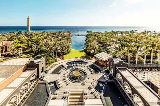 Udsigt over en hotelhave med palmer, en stor pool og hav i baggrunden. (AI-genereret tekst)