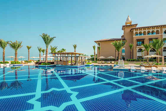 En swimmingpool med liggestole ved kanten, omgivet af palmer og en bygning i baggrunden. (AI-genereret tekst)