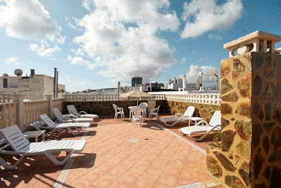 Liggestole og et bord med stole på en terrasse med udsigt over bygninger. (AI-genereret tekst)